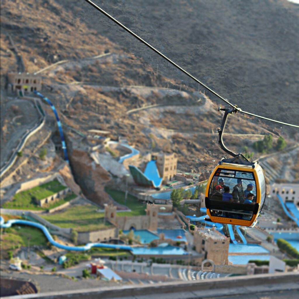لقطة سياحية احترافية لتلفريك الهدا في الطائف، تظهر عربات التلفريك وهي معلّقة فوق المنحدرات الجبلية وسط جبال الحجاز، مع إطلالة واسعة على الوديان والطبيعة المحيطة. تكون الصورة بإضاءة طبيعية ناعمة وأجواء صافية، مع إبراز ارتفاع التلفريك وجمال التضاريس الجبلية، بأسلوب واقعي وفاخر يعكس تجربة المغامرة والاستمتاع بالطبيعة.